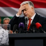 A defeated politician stands at a podium with press microphones and a ballot box in the foreground, Hungarian flag behind him — representing Viktor Orbán's election defeat in Hungary April 2026.
